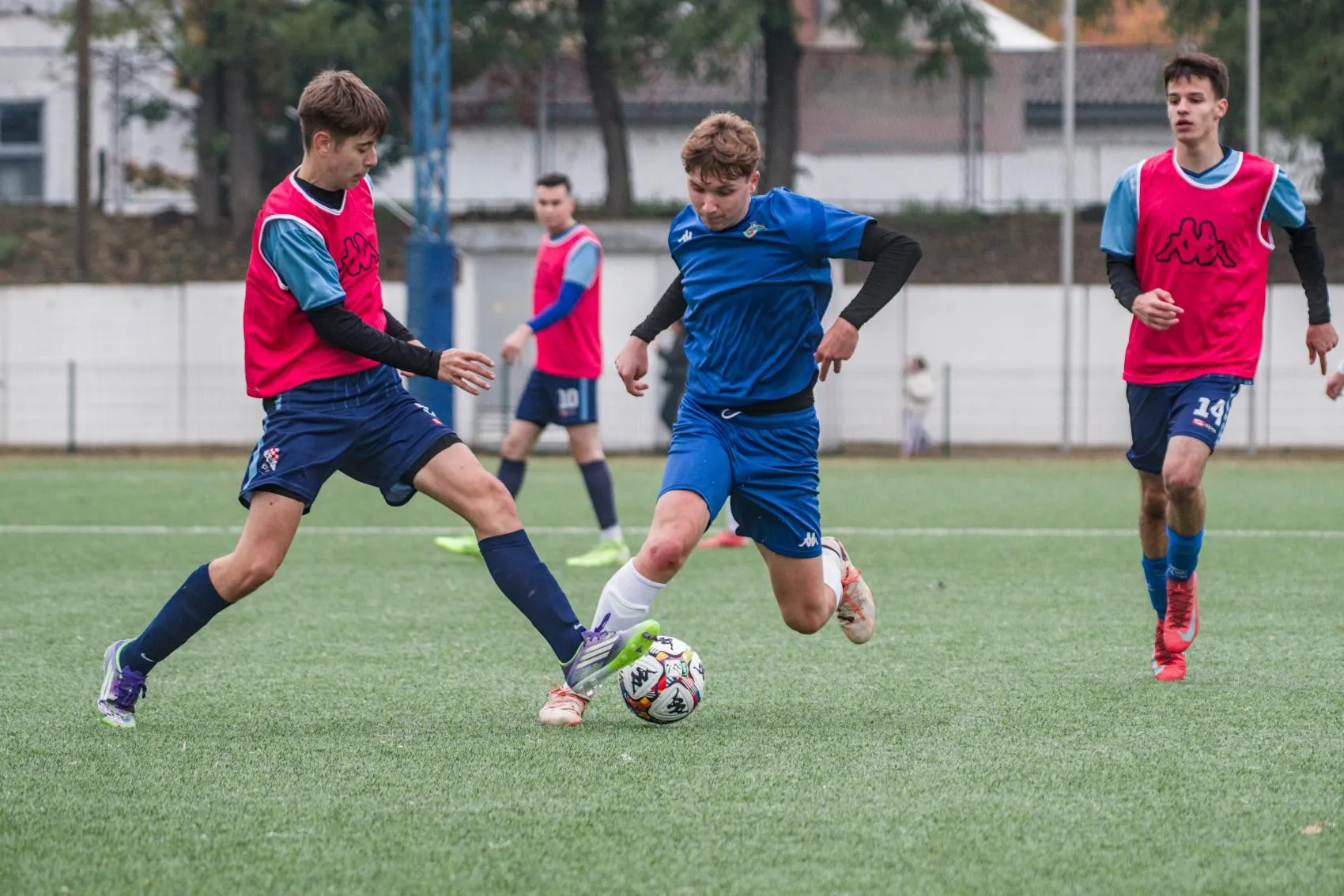 Mladi nogometaši u plavim i crvenim dresovima za trening natječu se za loptu na zelenom terenu tijekom treninga.